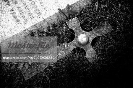 A black and white photograph of a cross, next to a tombstone.