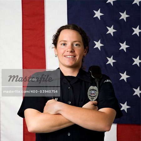 Portrait of mid adult Caucasian policewoman standing with arms crossed with American flag as backdrop smiling at viewer.