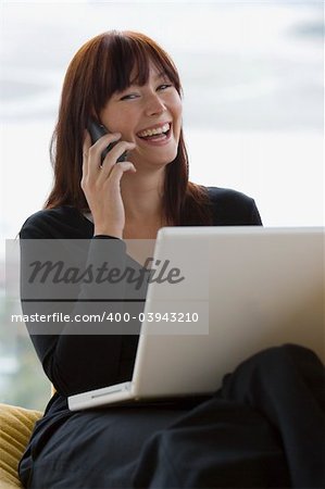 A young woman laughing while on a mobile phone and using a laptop computer