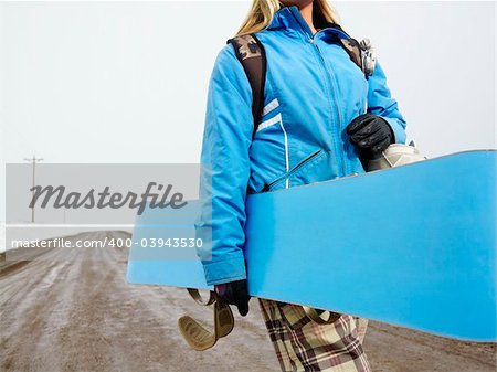 Young woman in winter clothes walking alone down muddy dirt road holding snowboard and boots.
