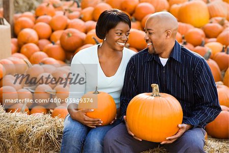 Happy smiling couple sitting on hay bales and holding pumpkins at outdoor market.