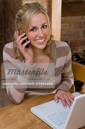 A beautiful young woman working from home on a laptop computer and talking on her mobile phone