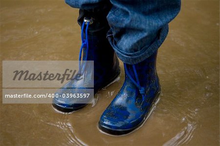 A little boy in blue boots standing in a muddy puddle