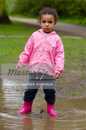 A little mixed race girl in pink coat and boots standing in a muddy puddle