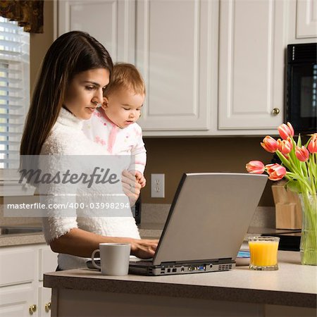 Mother holding baby  and typing on laptop computer in kitchen.