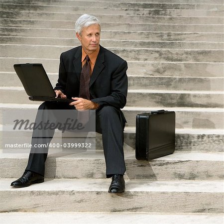 Caucasian middle aged businessman sitting on steps outdoors with laptop and briefcase.