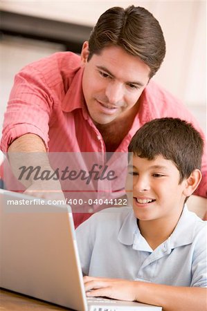 Man helping young boy in kitchen with laptop smiling