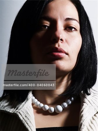 A high-key portrait about an attractive lady with black hair who is looking ahead and she has a glamorous look. She is wearing a white coat, a black dress and a white necklace.