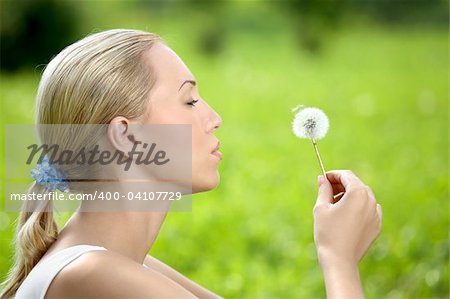The blonde sits in park on a grass with a dandelion in hands
