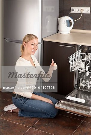 The young joyful woman sits near to the dishwasher on kitchen