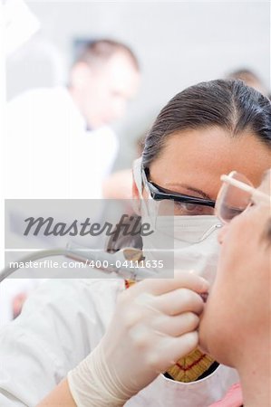 A young dentist treating an old woman's teeth whit the turbine, a doctor in the background - part of a series.