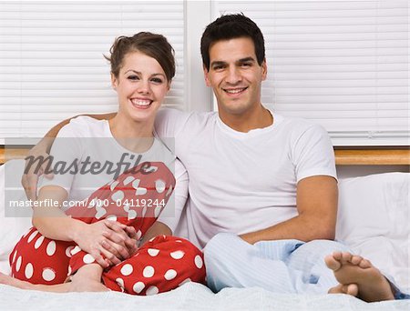 An attractive couple smiling and sitting in bed together.  Horizontally framed shot.