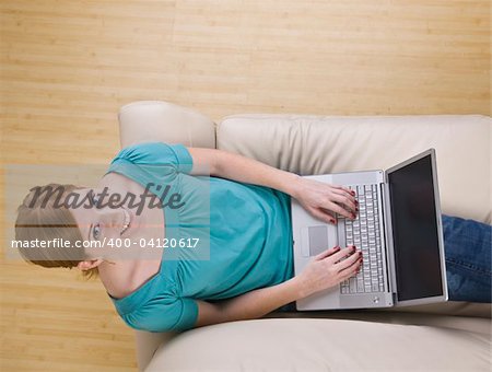 A view from above of a woman sitting on a couch and using a laptop computer.  She is looking up at the camera and is smiling. Horizontally framed photo.