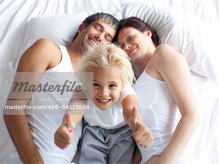 Happy little girl on bed with thumbs up smiling at the camera and her parents