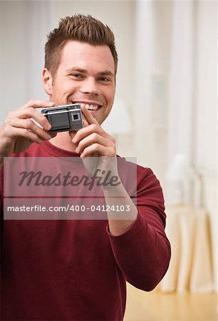 A young man is holding up a digital camera and smiling at the camera. Vertically framed shot.