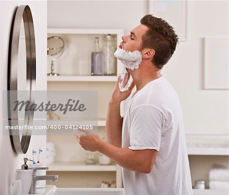 A man is shaving his face in the mirror.  He is looking away from the camera.  Horizontally framed shot.