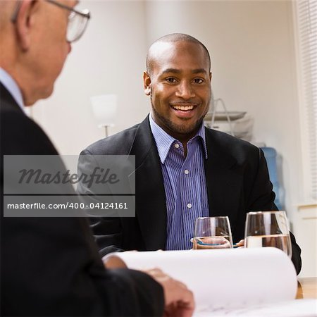 Two businessmen are in a meeting at an office.  The younger man is smiling at the camera.  Square framed shot.