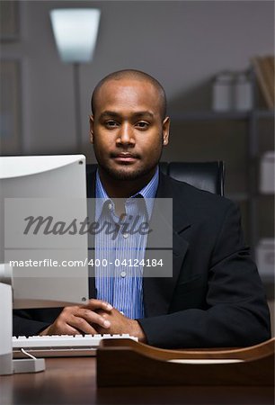 A young businessman is working on his computer at the office.  He is looking at the camera.  Vertically framed shot.