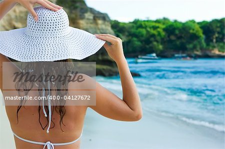 Beautiful brunette with long hair in white hat stands on the sand ocean beach on Bali in Indonesia