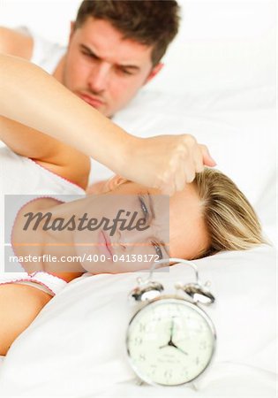 Attractive man and angry woman in bed looking at the alarm clock going off