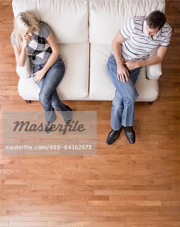 Man and a woman sit distantly on the ends of a cream colored love seat. Vertical shot.