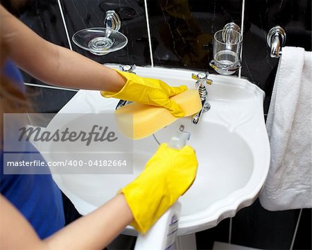 Young woman cleaning a bathroom's sink with a sponge and detergent spray