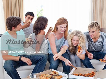 Six teenagers eating pizza in the living-room on the sofa