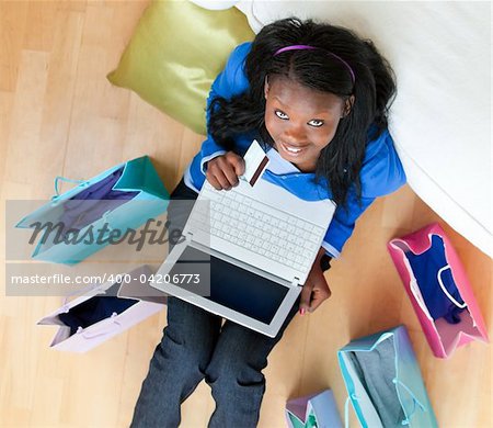 Cheerful afro-american teenager using a laptop sitting between shopping bags on the floor in the living room