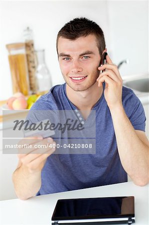 Attractive young man talking on phone holding a card looking at the camera in the kitchen