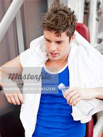 Tired man with a towel and a bottle of water sitting on a bench press in a fitness center