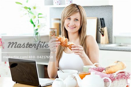 Smiling woman having breakfast in front of the laptop in the kitchen at home