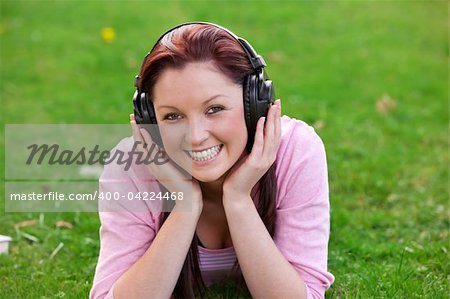 Delighted young woman listening to music with headphones lying on the grass in a park