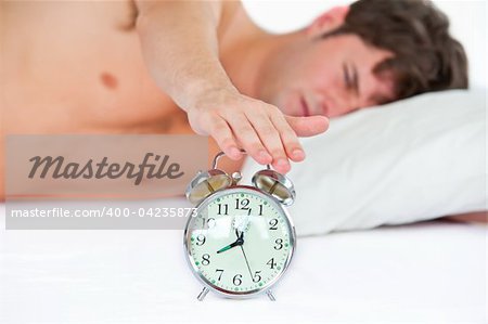 Asleep young man stopping his ringing alarm clock in the bedroom at home