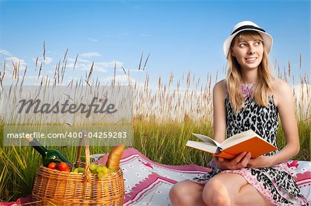 beautiful young woman having picnic on meadow, reading book and smiling. Looking away from camera, blue cloudy sky in background