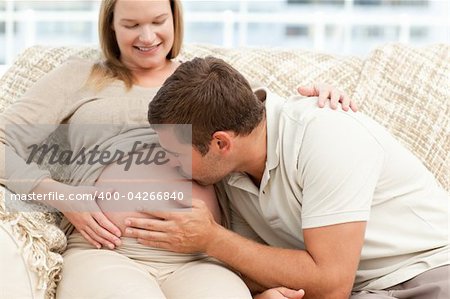 Future dad kissing the belly of his wife sitting on the sofa in the living room