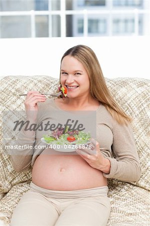 Happy pregnant woman enjoying a salad sitting on the sofa at home