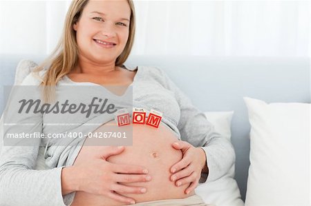 Blond pregnant woman with mom letters on her belly lying on a bed at home