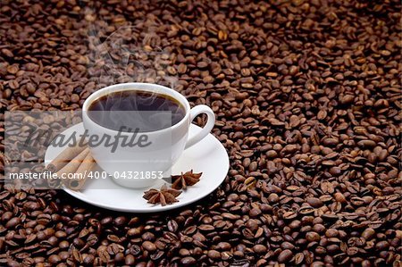 White cup of hot coffee on coffee beans with three sticks of cinnamon and star anise fruits