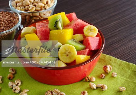 Fresh fruit salad made of banana, kiwi, watermelon and mango pieces in orange bowl with cereals (puffed wheat and puffed chocolate quinoa) (Selective Focus, Focus on the front of the bowl and the fruits in the front)