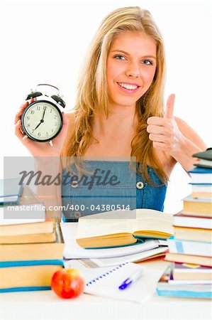 Smiling teen girl with alarm clock sitting at table and showing thumbs up gesture