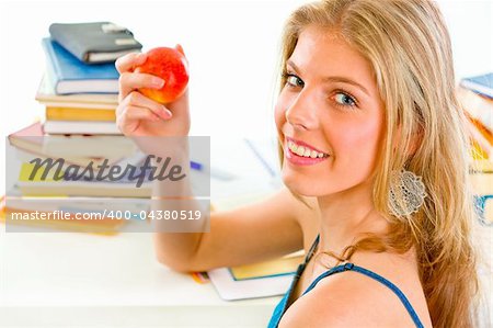 Smiling teengirl sitting at desk with books and holding apple in hand