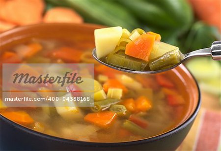 Fresh vegetable soup on spoon made of green bean, pea, carrot, potato, red bell pepper, tomato and leek in black bowl with ingredients in the back (Selective Focus, Focus on the carrot, leek and green bean on the spoon)