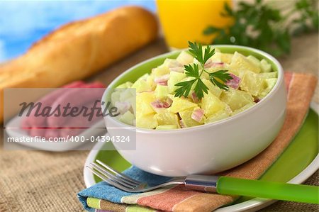 Potato salad made of cooked potatoes, red onions and cucumber, seasoned with a mayonnaise dressing and garnished with a parsley leaf with sausages, baguette and orange juice in the back (Selective Focus, Focus on the front of the salad and the leaf)