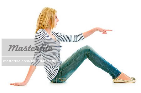 Beautiful teen girl sitting on floor and pointing finger at copy-space isolated on white