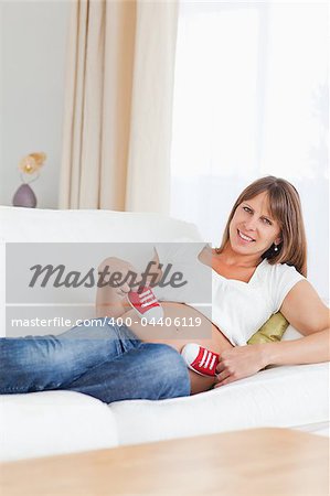 Cute pregnant female playing with red baby shoes while lying on a sofa in her living room