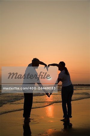 Mid-adult couple making heart shape with arms on beach at sunset.