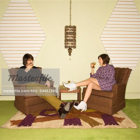 Caucasian mid-adult man and woman wearing vintage clothing seated in brown retro chairs smiling and drinking.