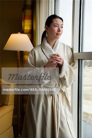 Taiwanese mid adult woman in bathrobe looking out window holding coffee cup.