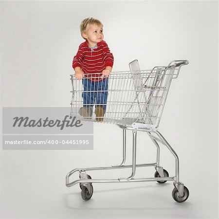 Studio portrait of Caucasian boy standing alone in empty shopping cart against white background.