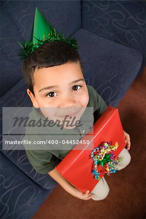 Hispanic boy wearing party hat holding birthday present looking up at viewer and smiling.